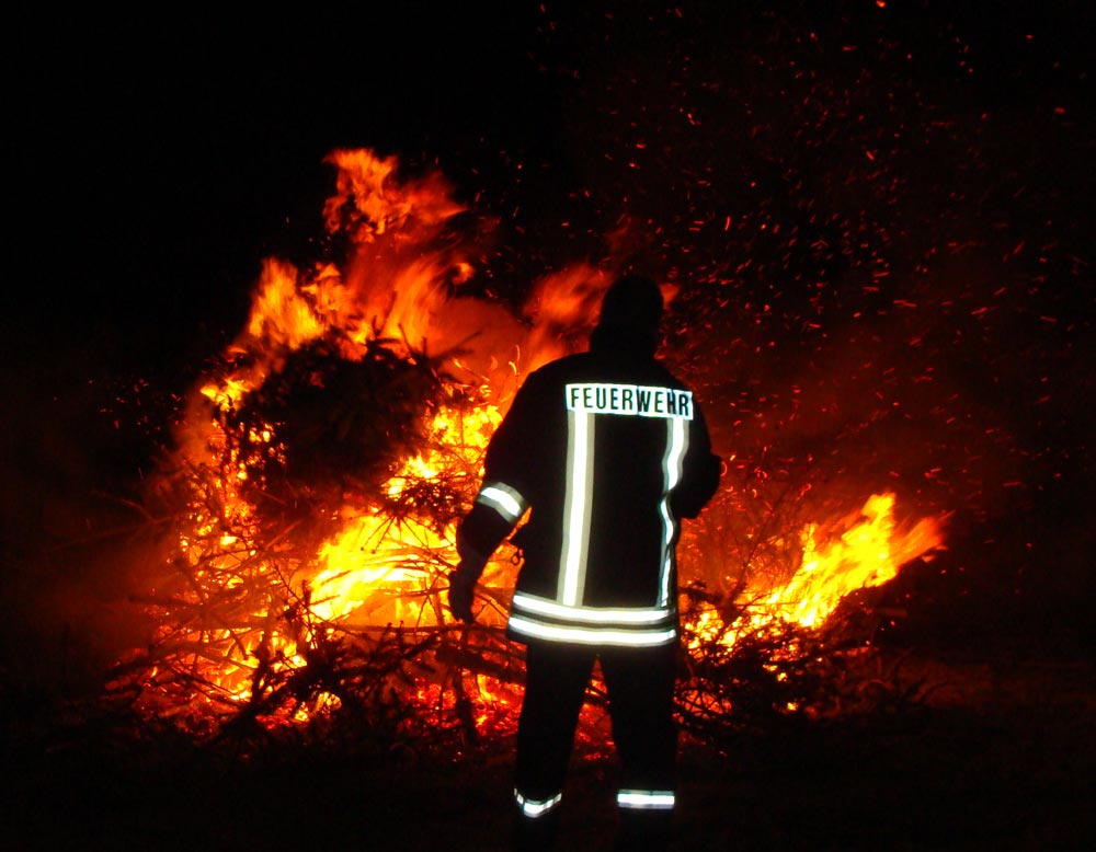 Ein Feuerwehrmann steht an einem brennenden Lagerfeuer aus Tannenbäumen.